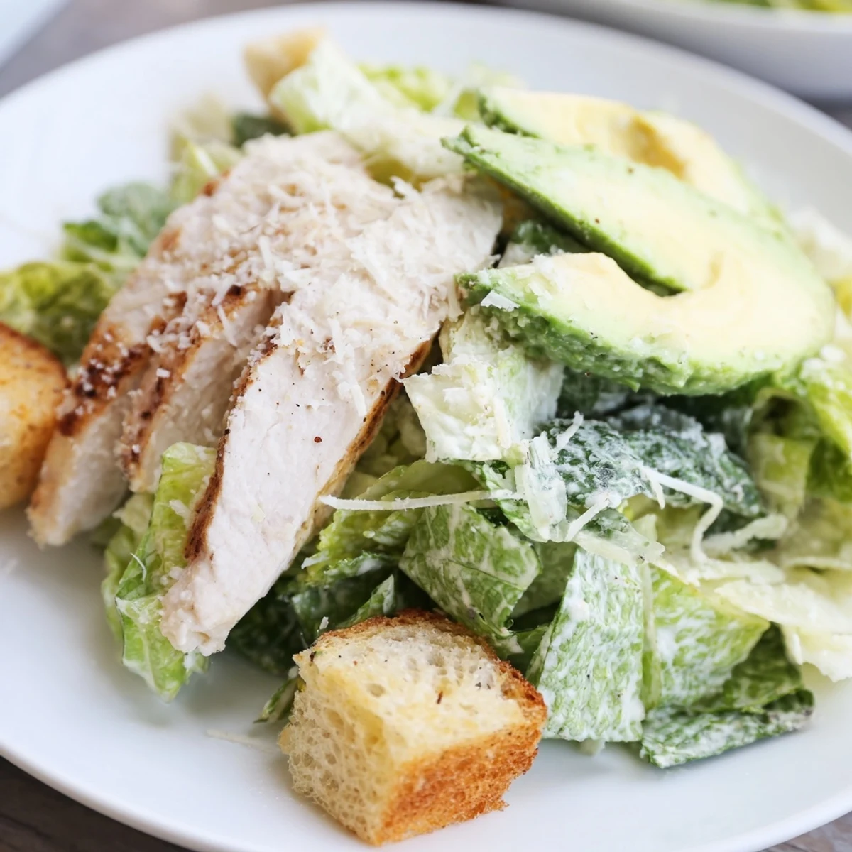 The bold, tangy flavors of Chicken Caesar Salad with Avocado shine in this plated dish, garnished with fresh parmesan and crunchy croutons for added texture.