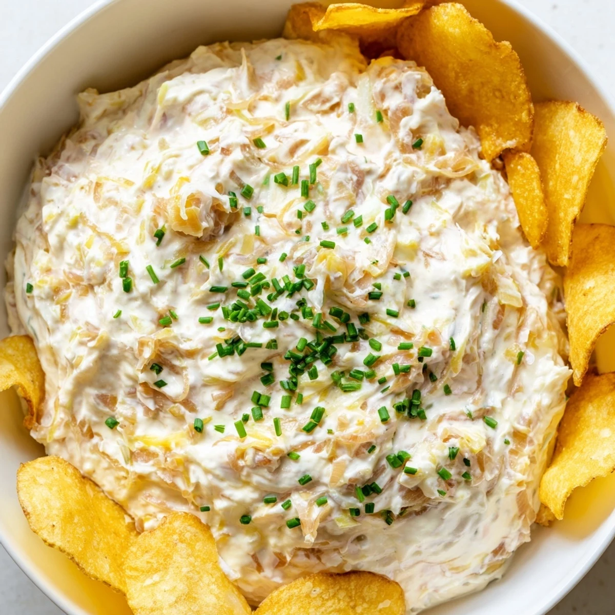 Golden caramelized onion dip topped with fresh chives, served alongside crunchy, thick-cut potato chips on a rustic plate.