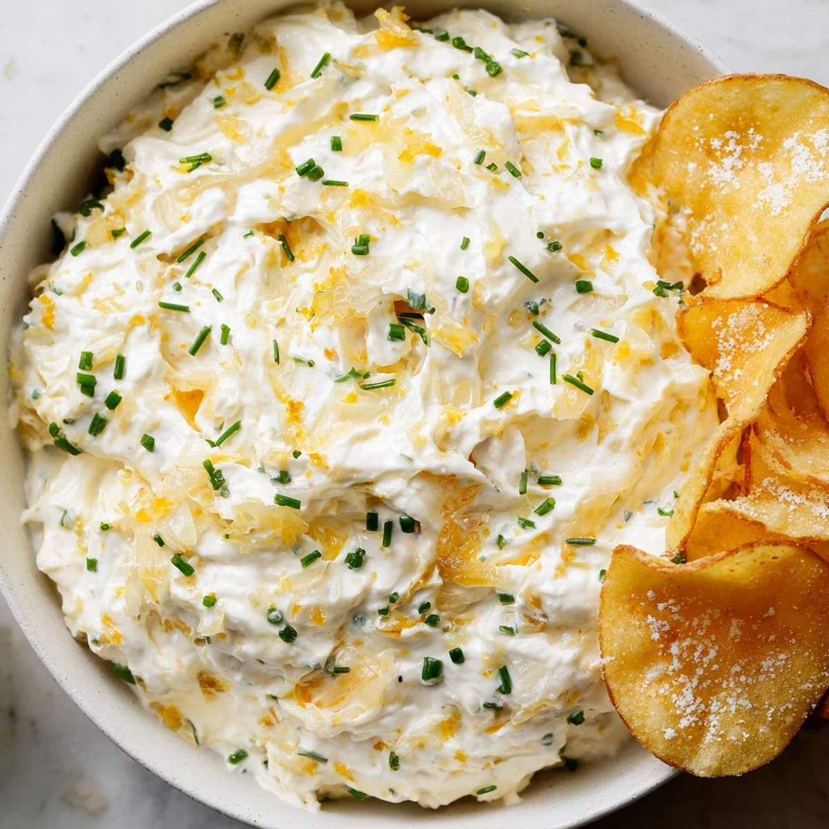 Golden caramelized onion dip in a rustic bowl sits beside homemade, crispy potato chips, perfect for snacking.
