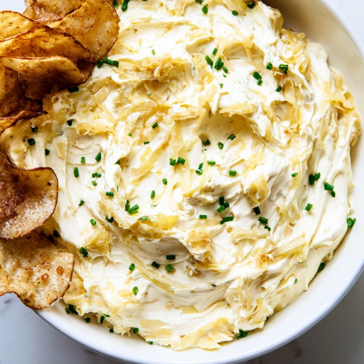 A close-up of creamy onion dip with chopped chives and a pile of golden, freshly fried potato chips.